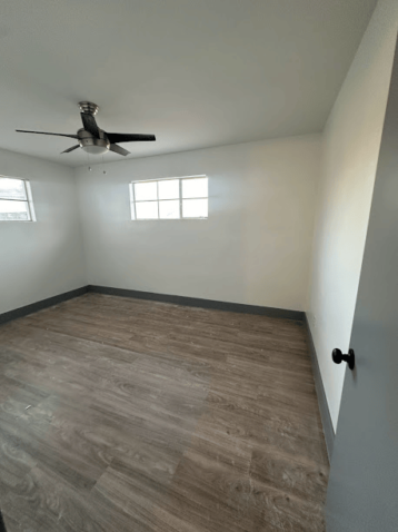 Empty room with ceiling fan, wood flooring, and two windows.