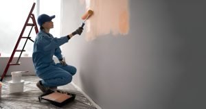 Female painter in overalls with cap and gloves painting a wall with paint roller, ladder and buckets on background
