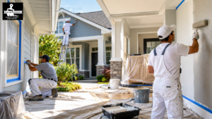 Painters in white overalls painting the exterior of a blue house with ladders and drop cloths.